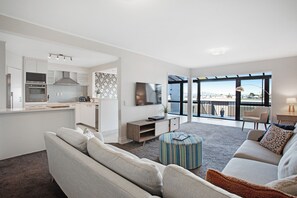 A modern living room with a sofa, ottoman, and a dining area with a table and chairs. There is a kitchen with white cabinets and a large window offering a view of the city. - Parnell Vista 2BR Apartment with Seaview (Auckland)