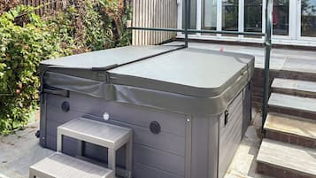 A spa hot tub with a small table and bench, located on a stone patio next to a wooden fence and steps leading to a glass door.