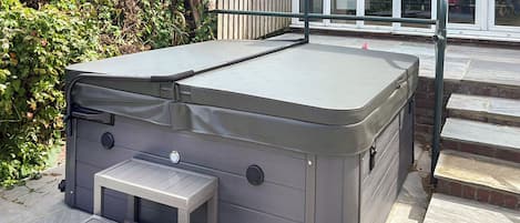Un jacuzzi de spa con una pequeña mesa y reposición, situado en un patio de piedra junto a una valla de madera y escalones que conducen a una puerta de cristal.