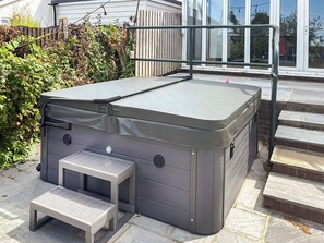 A spa hot tub with a small table and bench, located on a stone patio next to a wooden fence and steps leading to a glass door.