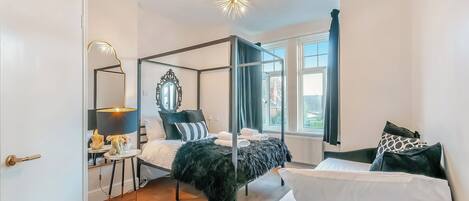 Un dormitorio con cama con dosel, un espejo redondo, una mesita de noche con lámpara y vistas al exterior a través de una ventana.