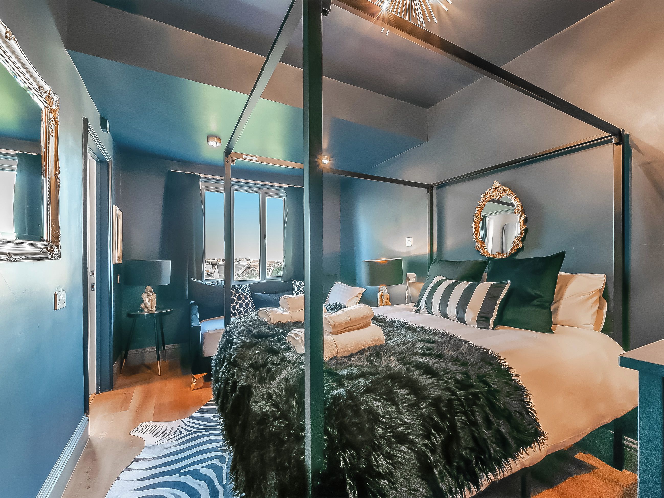 A modern bedroom with a four-poster bed, a large mirror, and a view of the city through the window.