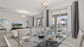 A modern dining area with a glass table, white chairs, and a chandelier. - Luxury 6BR Escape – Pool, Water Slides & Lazy River Near Disney! (Four Corners)