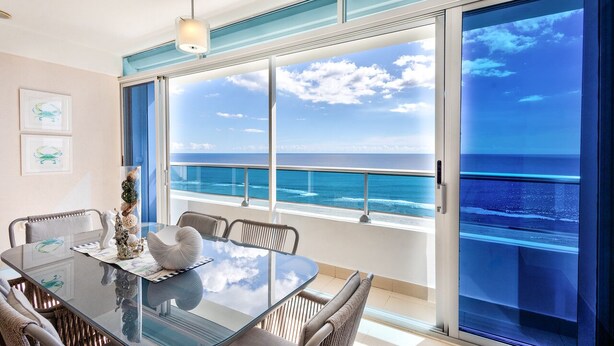 A dining area with a glass table and chairs, large windows with a view of the ocean, and a ceiling light fixture. - Beautiful 2-bedroom condo in fabulous Guayacanes with WiFi, AC (Guayacanes)