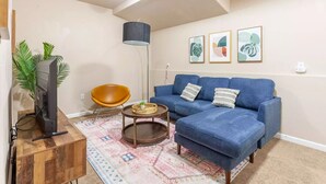 Uma sala de estar com um sofá azul, um móvel de televisão em madeira, um abajur, um tapete estampado e obras de arte emolduradas na parede.