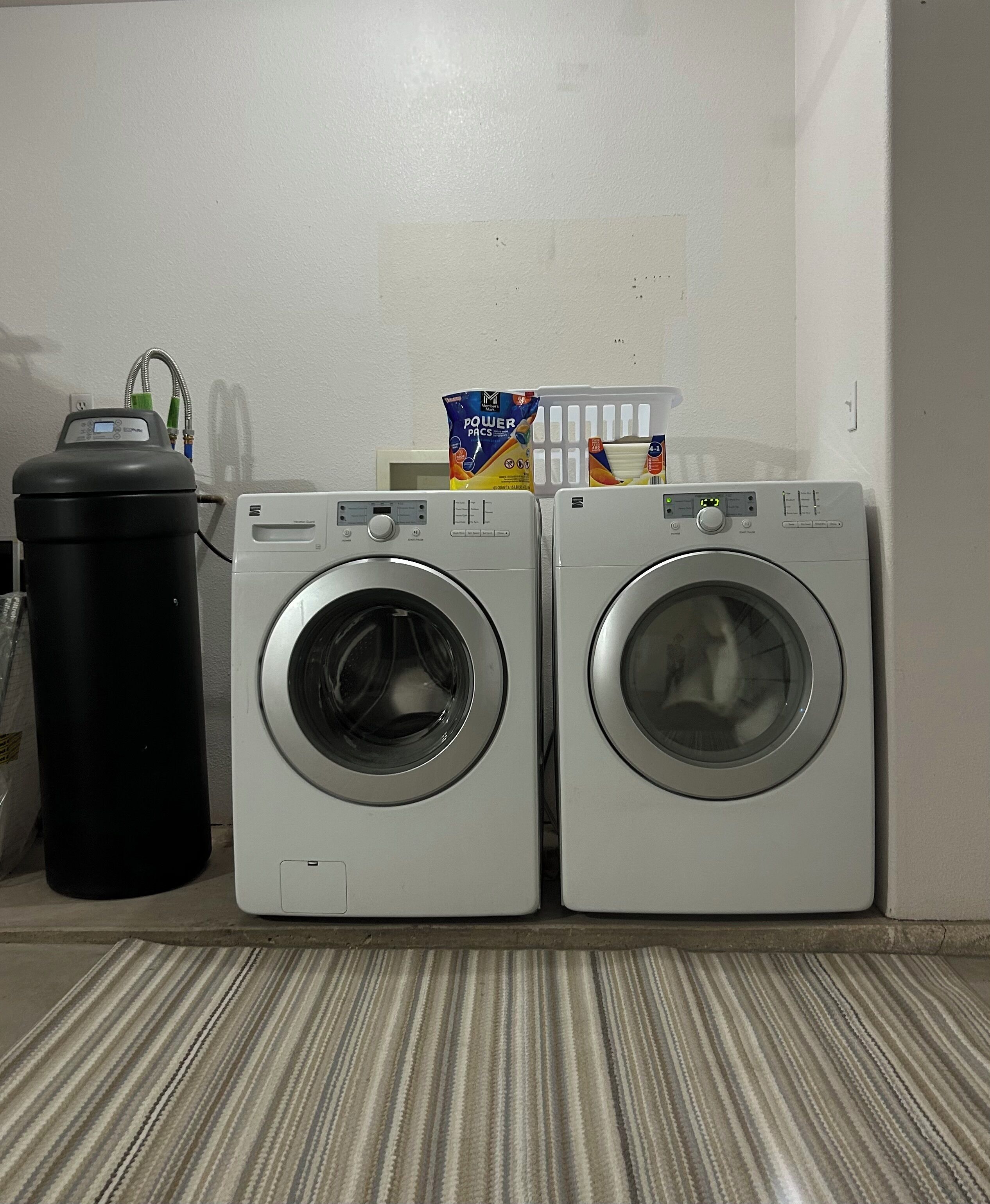 Two front-loading washing machines next to a black trash can and a laundry detergent box on a striped floor.