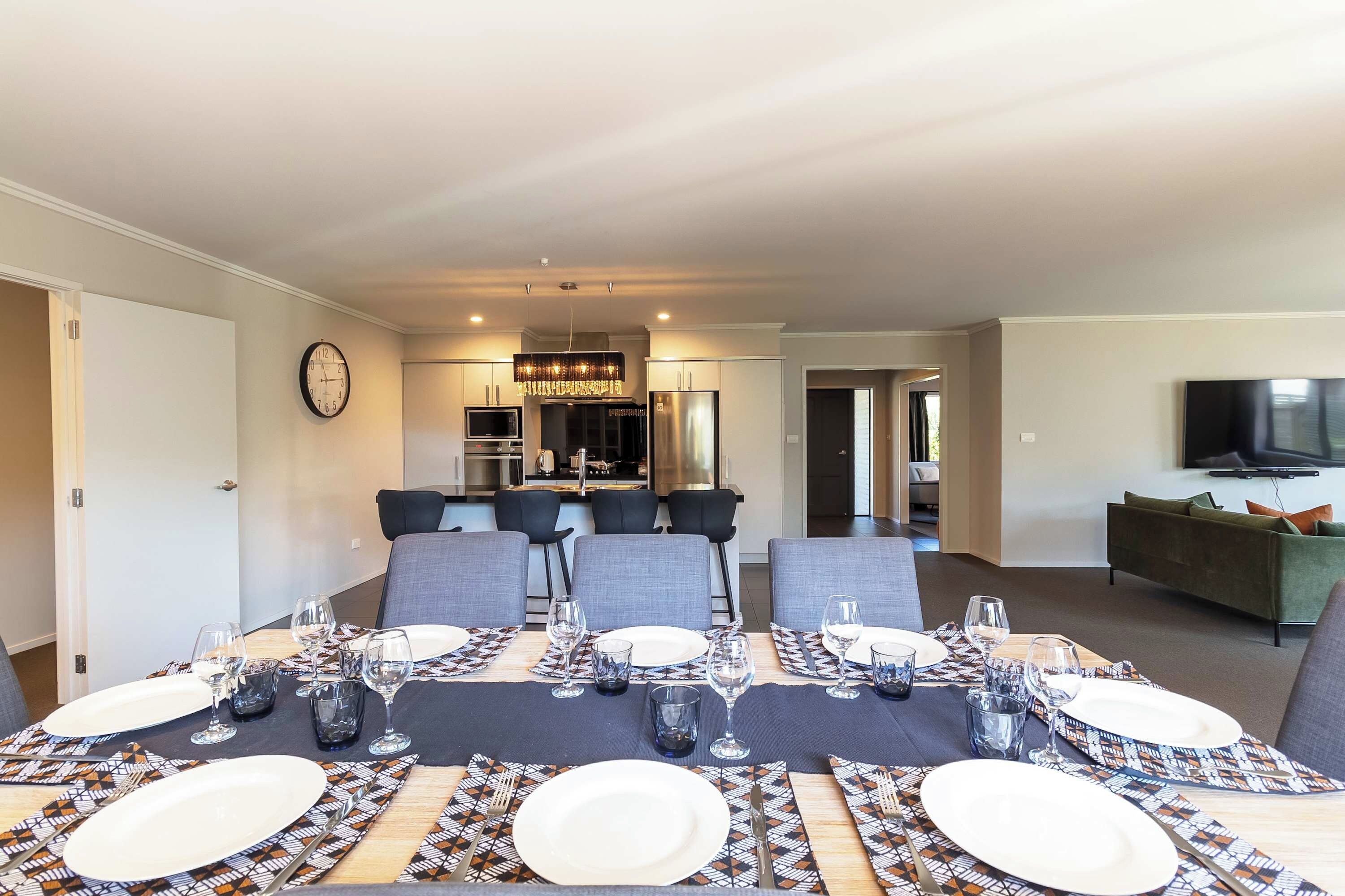 A dining area with a table set for four, a kitchen in the background, and a living room with a sofa and TV.