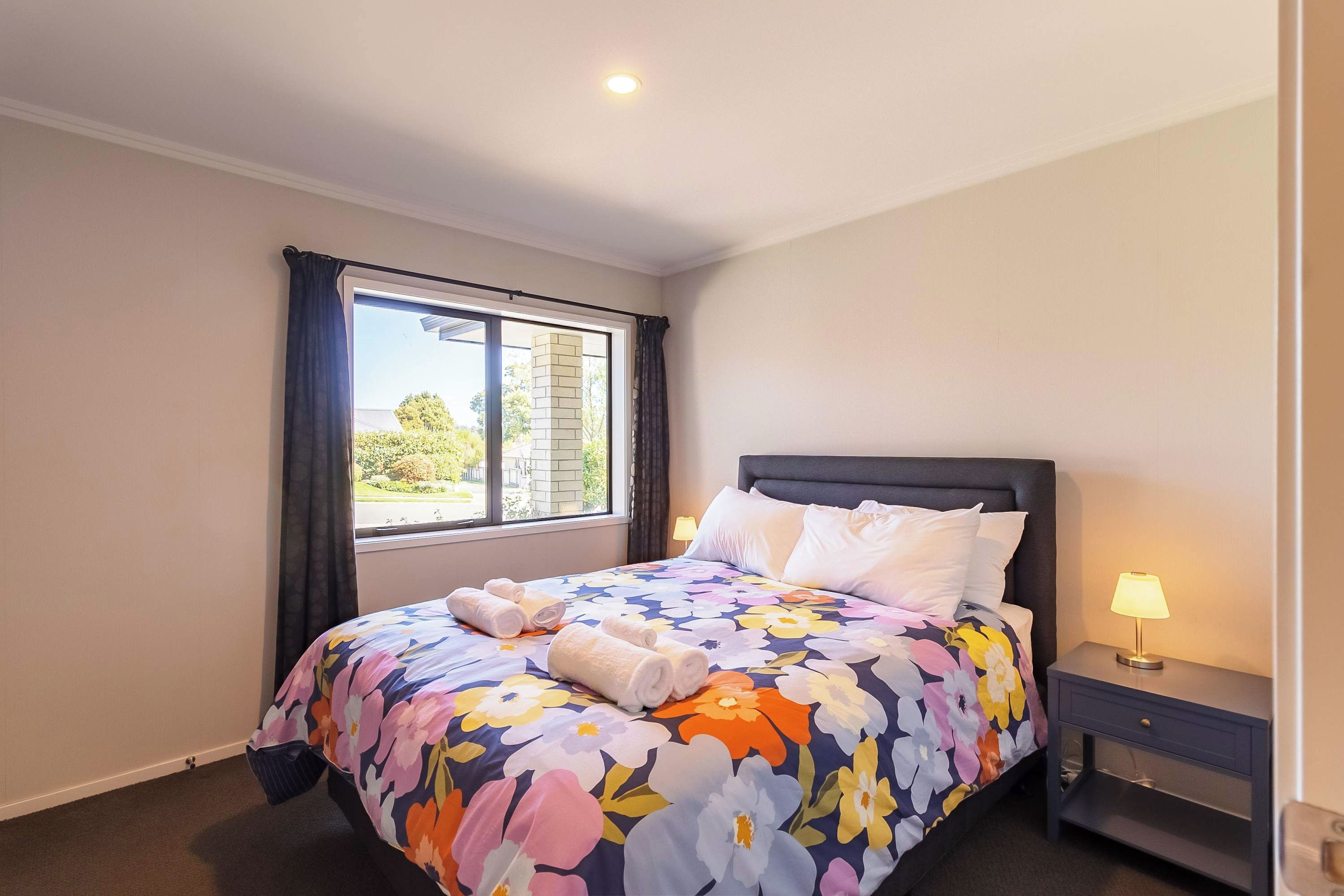 A neatly made bed with a floral bedspread, a bedside lamp, and a window with curtains.