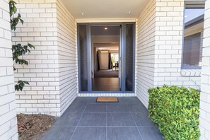 A modern building entrance with a dark tiled floor, white brick walls, and a green hedge.