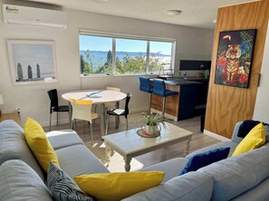 A modern living room with a grey sofa, yellow pillows, a round dining table with chairs, a kitchen area with a blue island, and a large window offering a view of trees and hills.