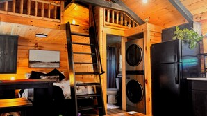 A compact cabin interior with a wooden interior, a bed with pillows, a table, a ladder, a washing machine, a refrigerator, and a window with a plant. - New Cabin for an Adventure (Peculiar)