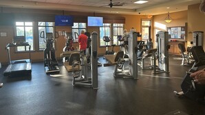 A gym with various exercise machines, a television displaying news, and large windows allowing natural light.