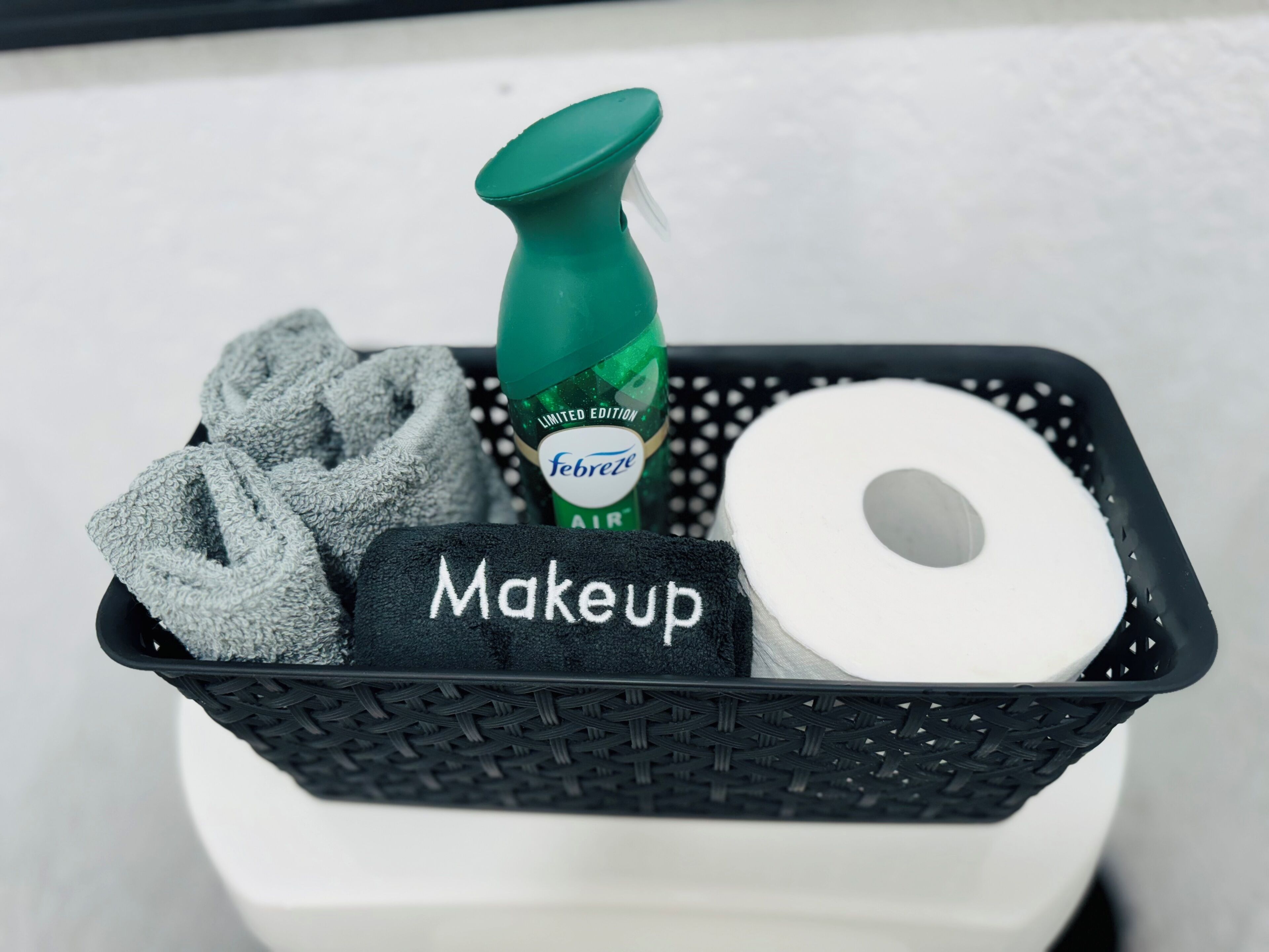 A black basket containing a green spray bottle labeled "Febreze Air," a roll of toilet paper, and a black towel with "Makeup" written on it.