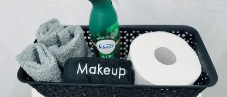 A black basket containing a green spray bottle labeled "Febreze Air," a roll of toilet paper, and a black towel with "Makeup" written on it.
