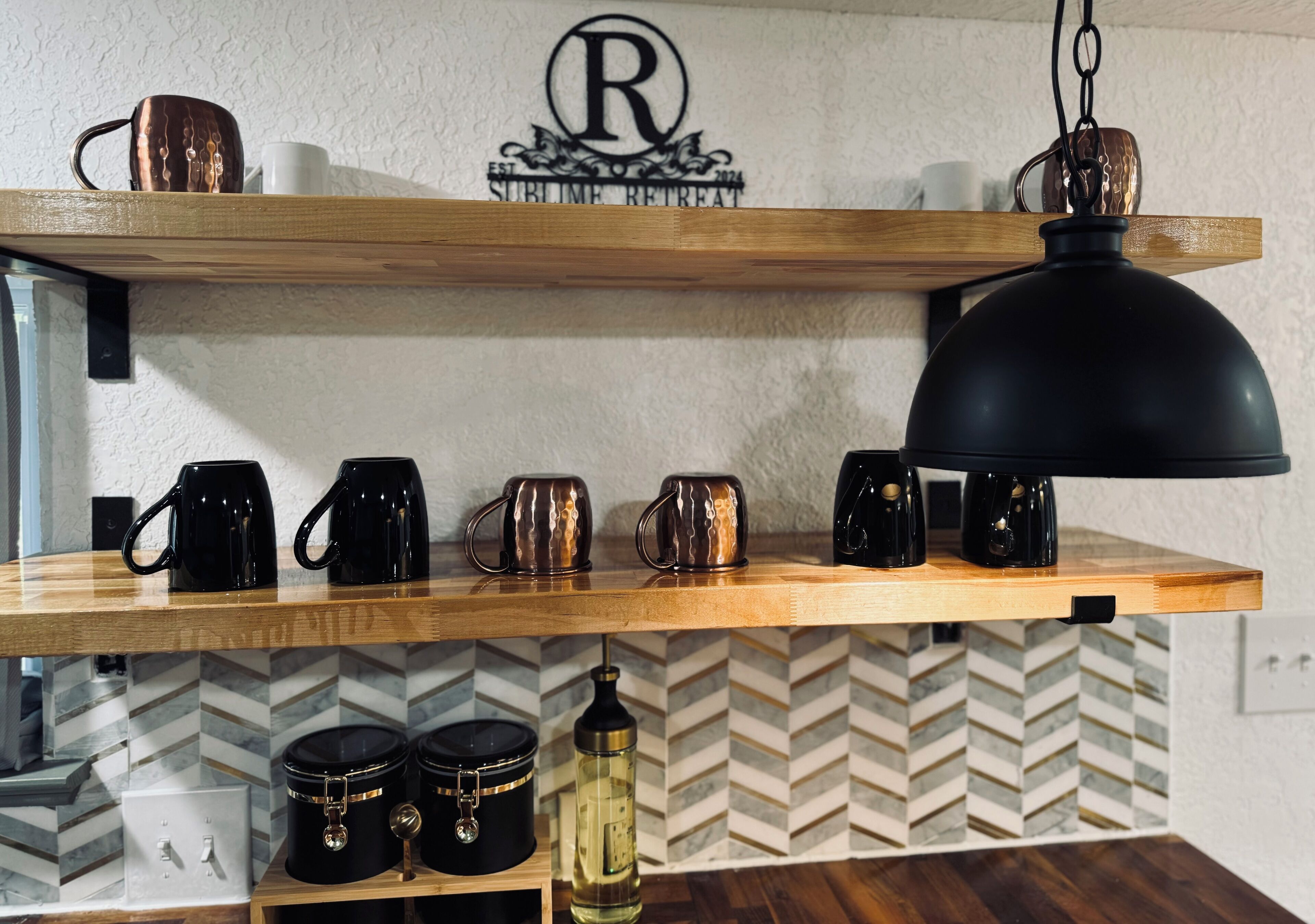 A kitchen with wooden shelves holding mugs and a coffee maker. There is a logo on the wall and a pendant light above the shelves.