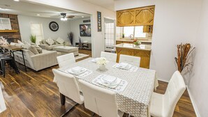 A dining area with a table set for four, surrounded by white chairs, in a room with a wooden floor and a kitchen area visible in the background. - Resort Pool & Spa Retreat with Game Room, Patio, Foosball & BBQ (North Richland Hills)