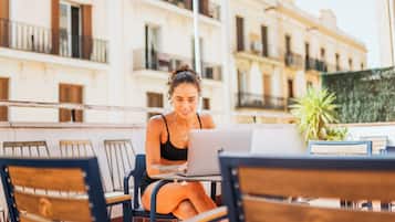 Una persona sentada a una mesa con una computadora portátil, en un espacio al aire libre con un edificio al fondo.