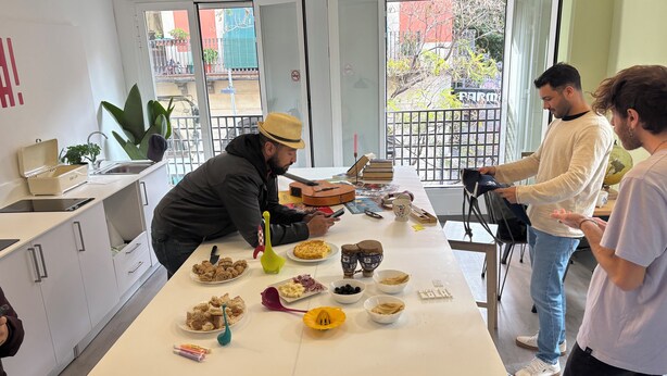 A kitchen area with a long table set for a meal, various food items, and kitchen utensils. - Hey Lola Hostel (Barcelona)