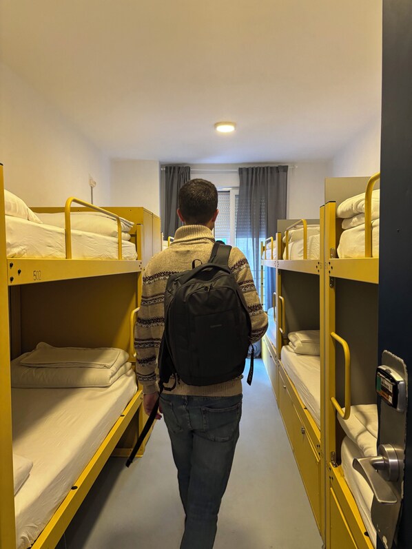 A narrow corridor with bunk beds on either side, a person walking towards a window with curtains, and a ceiling light. - Hey Lola Hostel (Barcelona)