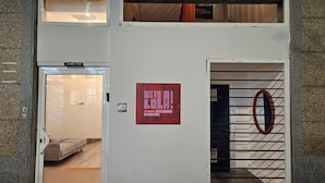 A hostel entrance with a red sign, a door with a circular emblem, and a glimpse of a room with a bed and a table. - Hey Lola Hostel (Barcelona)