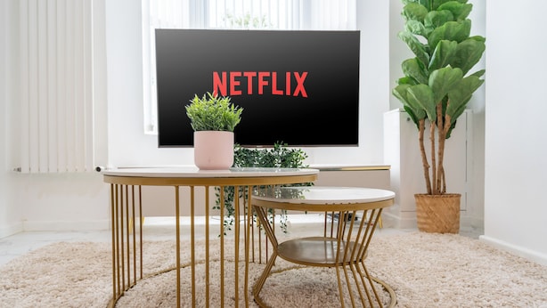 A room with a large flat-screen TV displaying the Netflix logo, a round gold table with a potted plant, and a tall green plant beside the TV.