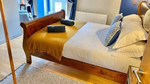 Ein hölzernes Bett mit gelber Decke, weißer Bettwäsche und dekorativen Kissen. Ein runder Spiegel hängt an der Wand, und eine Lampe steht auf dem Nachttisch.