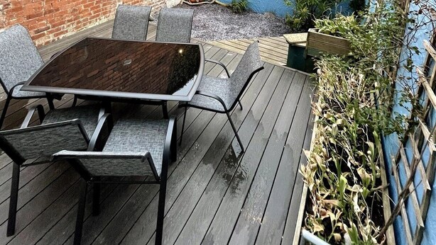 Eine Holzterrasse mit Tisch und Stühlen, eine Backsteinmauer und ein blaues Gebäude im Hintergrund.
