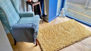 Ein Zimmer mit einem blauen Sessel, einem kleinen Holztisch mit einer Salzlampen und einer Statue, sowie einem großen, beigen Teppich auf Holzdielen.