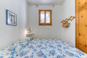 A bedroom with a bed, a window, a wooden wardrobe, a small table, and a framed picture on the wall.