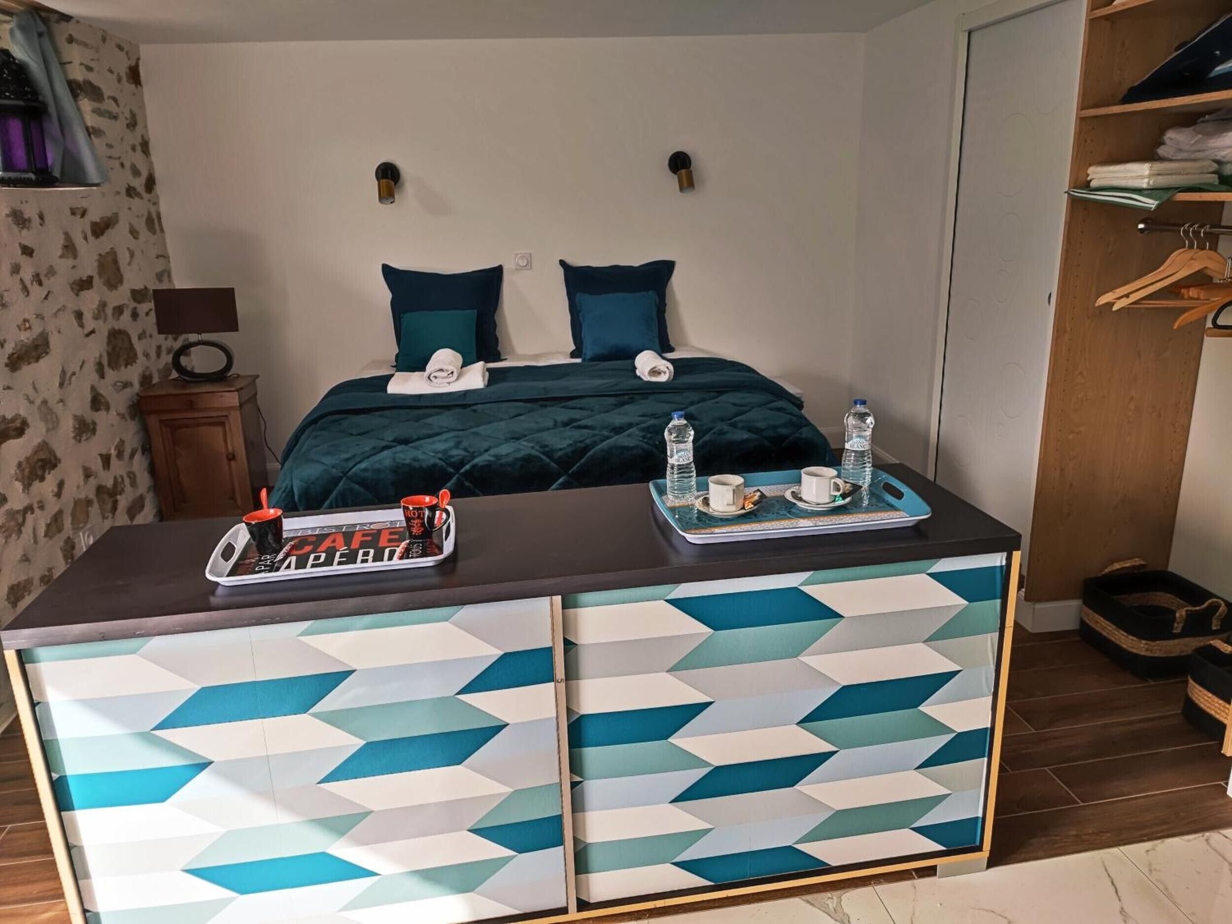 A hotel room with a bed, bedside tables, a desk with a tray of water bottles and cups, and a patterned cabinet.