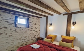 Une chambre avec des poutres apparentes, un mur en pierre, une fenêtre avec des rideaux bleus et deux lampes fixées au mur.