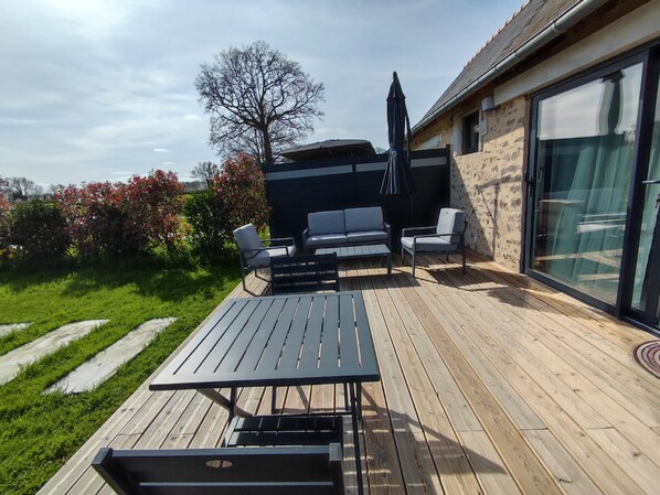 Une terrasse en bois avec des meubles d’extérieur, une porte vitrée et une vue sur un jardin.