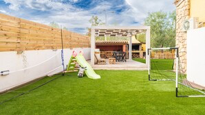 Ein Garten mit Kunstgrasanlage, einem Spielbereich für Kinder mit Rutsche, einer Terrasse mit Gartenmöbeln und einer Steinmauer.