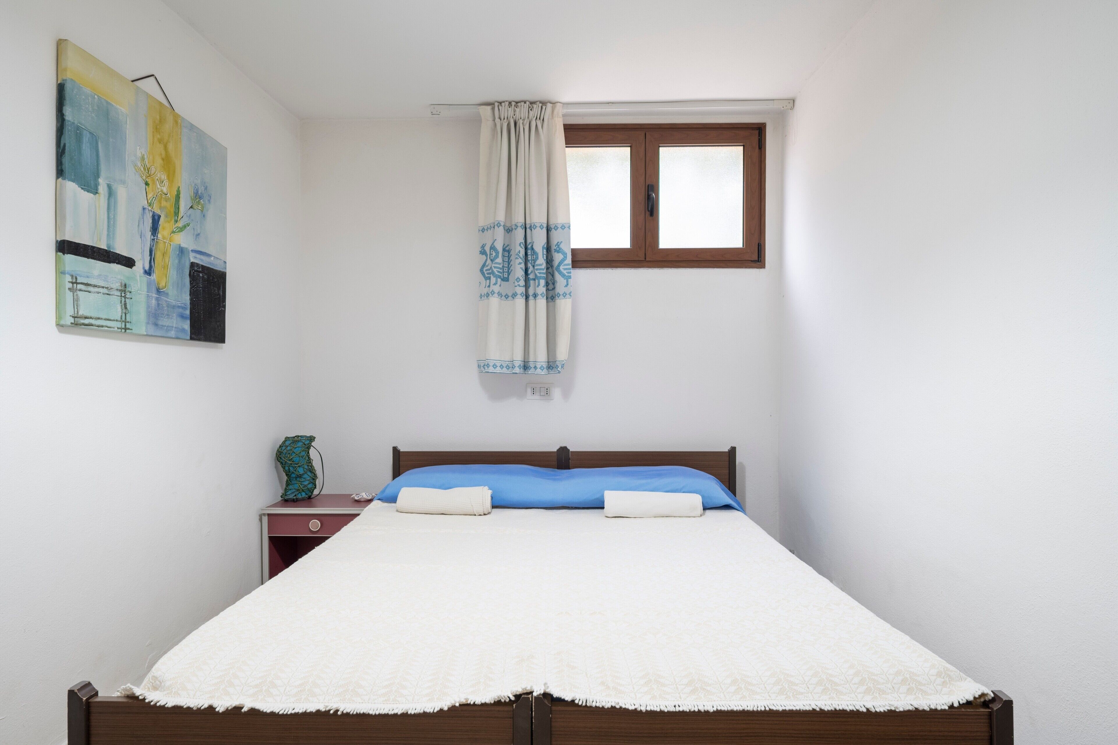Une chambre à coucher avec un lit, une table de chevet, une fenêtre avec des rideaux et un tableau accroché au mur.