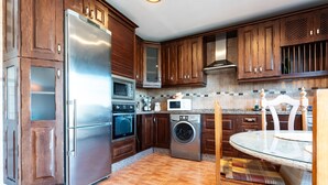 A kitchen with wooden cabinets, stainless steel appliances, a washing machine, and a dining area with a glass table. - Holiday Home 'The Palm House' with Mountain View, Wi-Fi and Air Conditioning (Las Lagunas de Mijas)