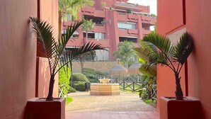 Un couloir donnant sur un bâtiment à la façade rouge, bordé de palmiers et agrémenté d’une fontaine.