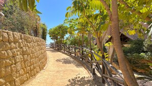 Un chemin pavé, bordé d’un côté par un mur de pierres et de l’autre par une clôture en bois, le tout entouré de verdure.
