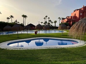 Una piscina envoltada per gespa, palmeres i un edifici amb façana vermella.