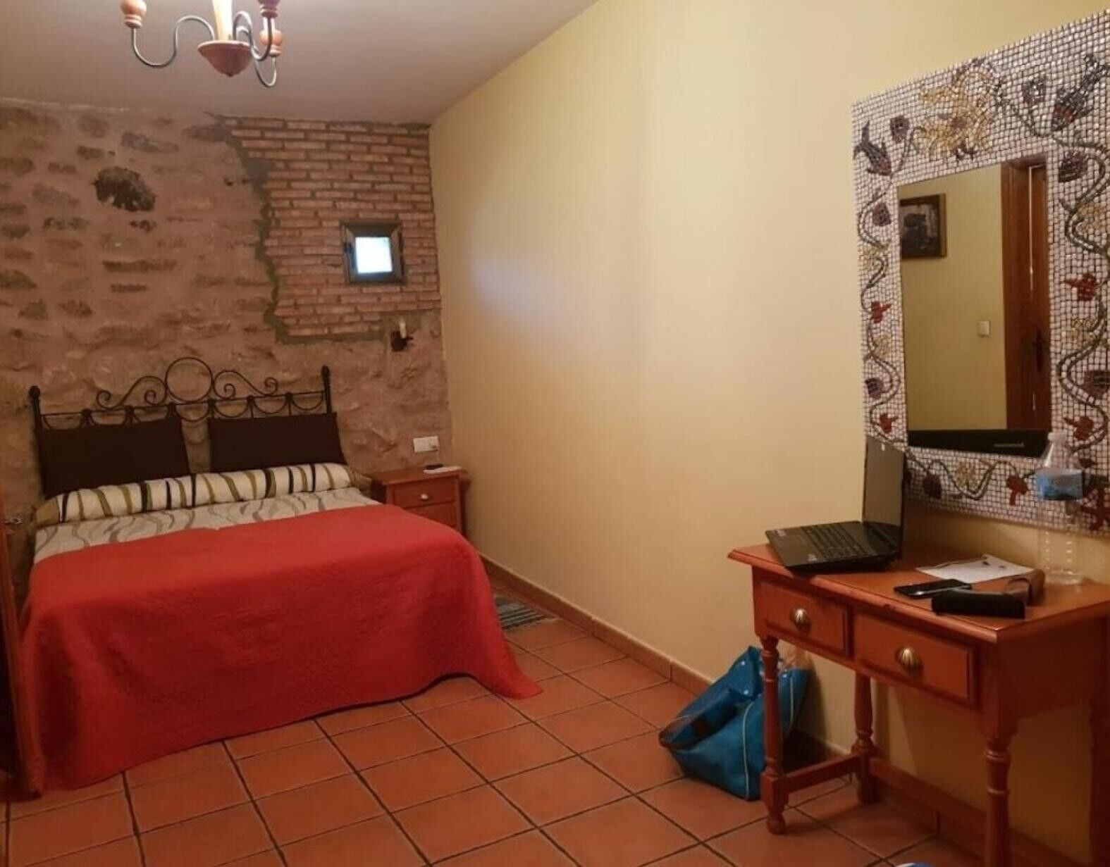 Une chambre avec un mur en briques, un lit recouvert d’une couverture rouge, une table de chevet en bois, un bureau avec un ordinateur portable et un miroir doté d’un cadre décoratif.