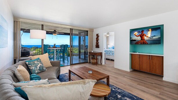 A modern living room with a balcony overlooking the ocean, a large flat-screen TV, and a cozy seating area.