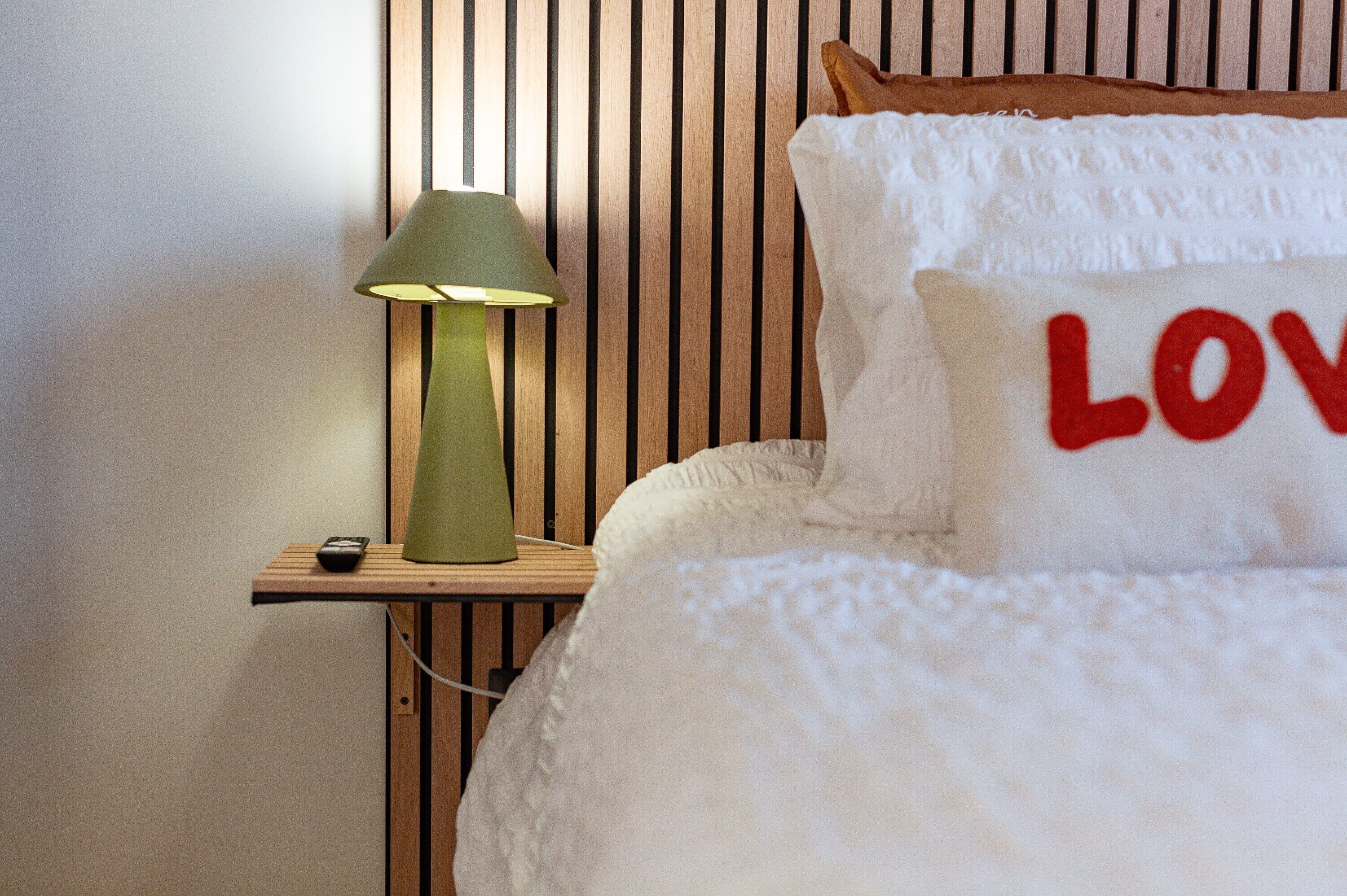 Ein Bett mit weißer Decke und Kissen, eines mit dem Wort 'LOVE' in roten Buchstaben, eine grüne Lampe auf einem Nachttisch aus Holz und eine Fernbedienung.