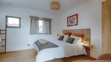 Een slaapkamer met een bed, een nachtkastje met een lamp, een raam met gordijnen, een lijst met een afbeelding aan de muur en een houten vloer.