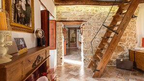 Un intérieur rustique avec un mur en pierre, un escalier en bois et une commode en bois surmontée d’une horloge.