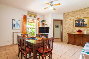 A dining area with a table, chairs, a ceiling fan, a TV, and a brick wall with a painting.