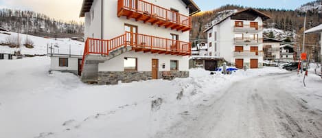 Un edificio de dos pisos con un suelo cubierto de nieve, una escalera que lleva a un balcón y un cielo despejado.