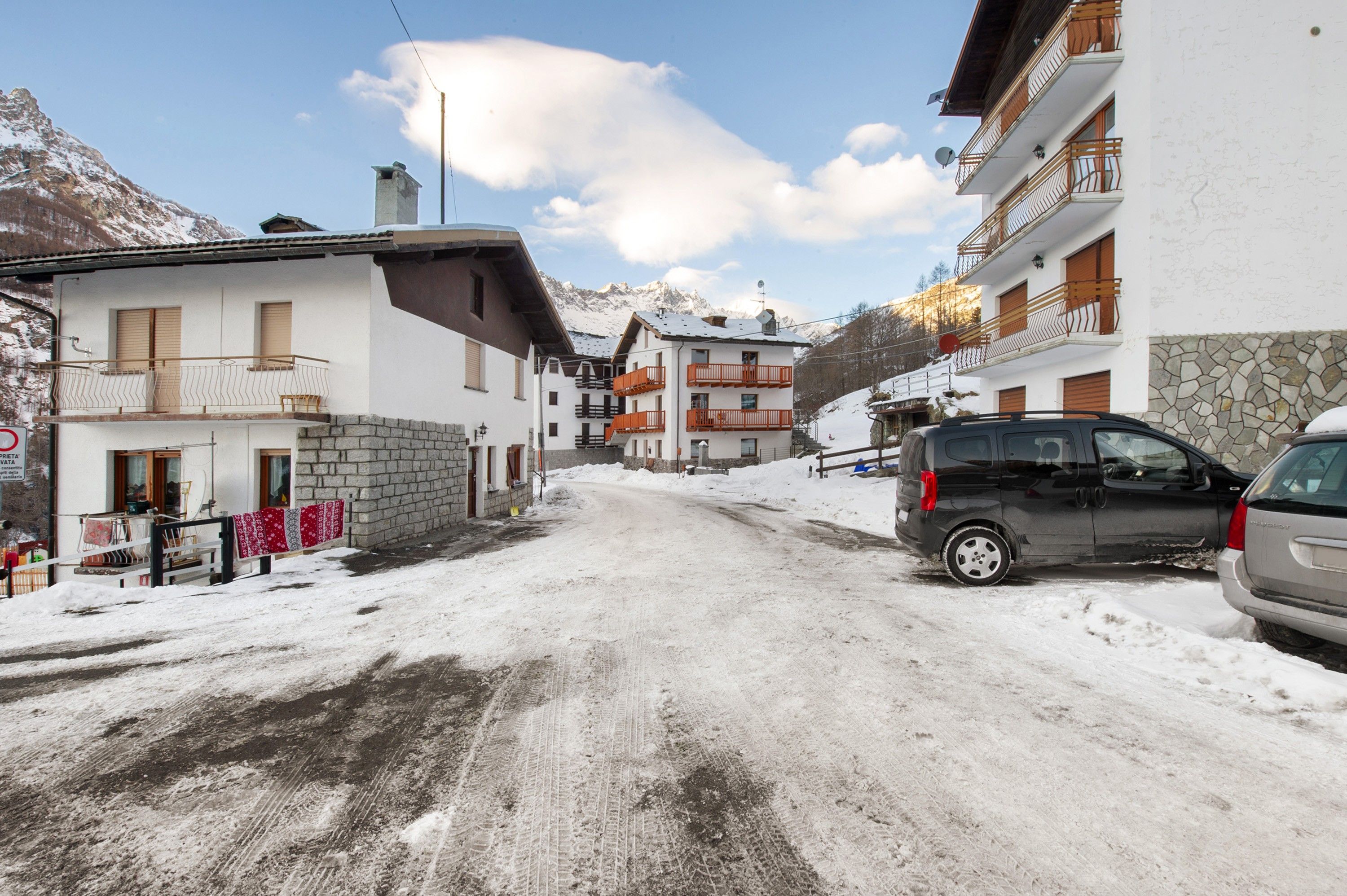 Une rue enneigée bordée d’immeubles, avec une voiture garée et une montagne en arrière-plan.