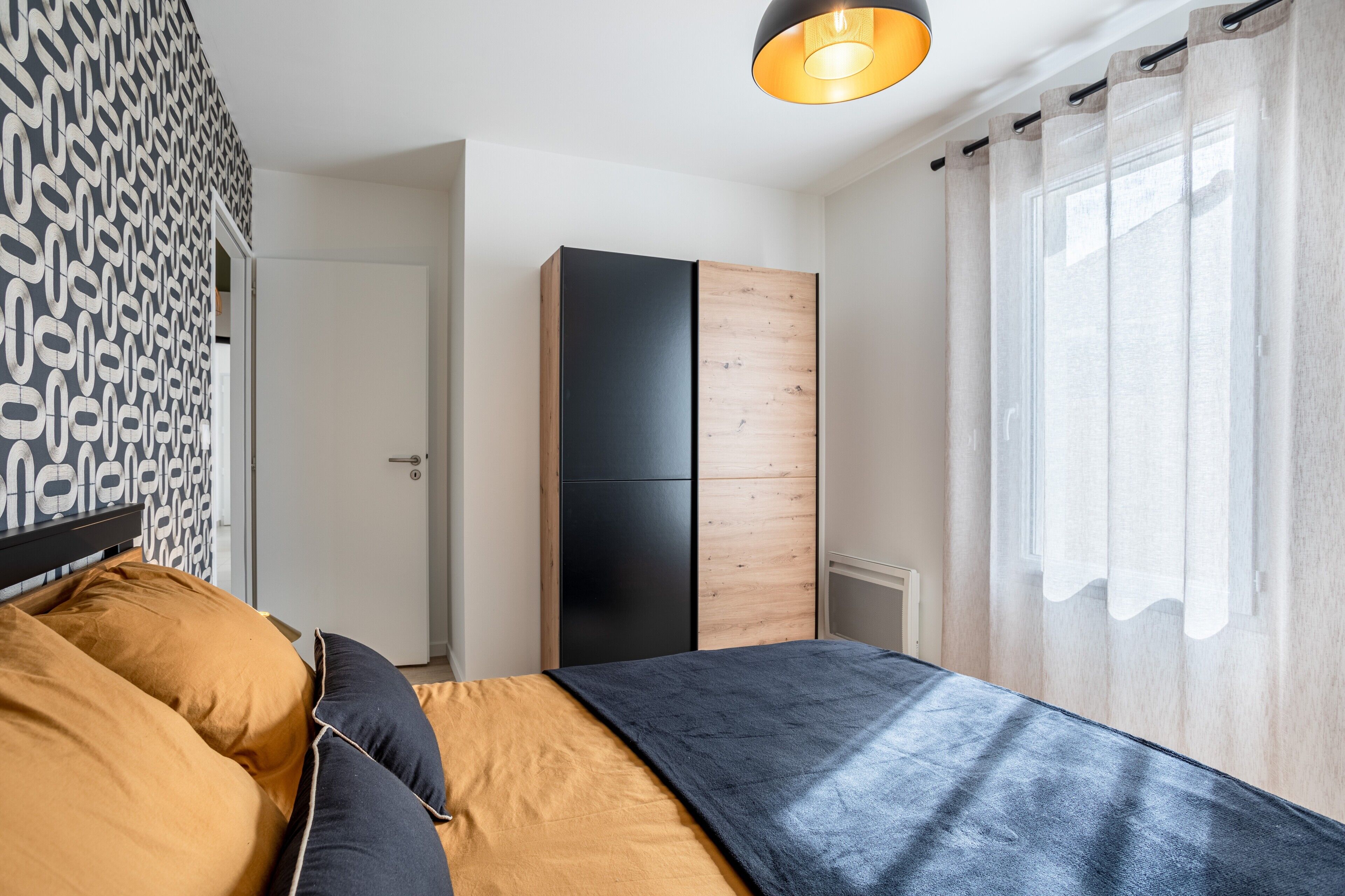 A bedroom with a bed, a wardrobe, a window with curtains, and a patterned wall.