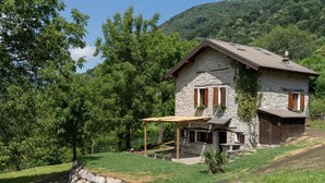 A stone house with a sloped roof, wooden shutters, and a covered patio area. - Chalet 'Nini's Home' with Terrace and Garden with BBQ (Schignano)