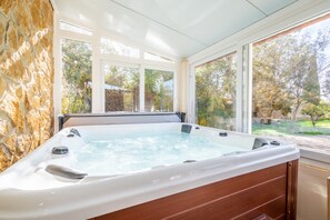 A hot tub with a stone wall backdrop, large windows, and a view of trees.