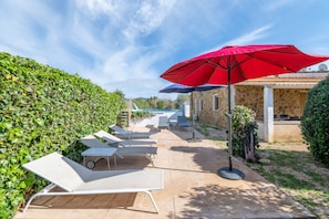 Eine Terrasse mit Liegestühlen, einem roten Sonnenschirm, einem Steinhaus und einer Hecke.